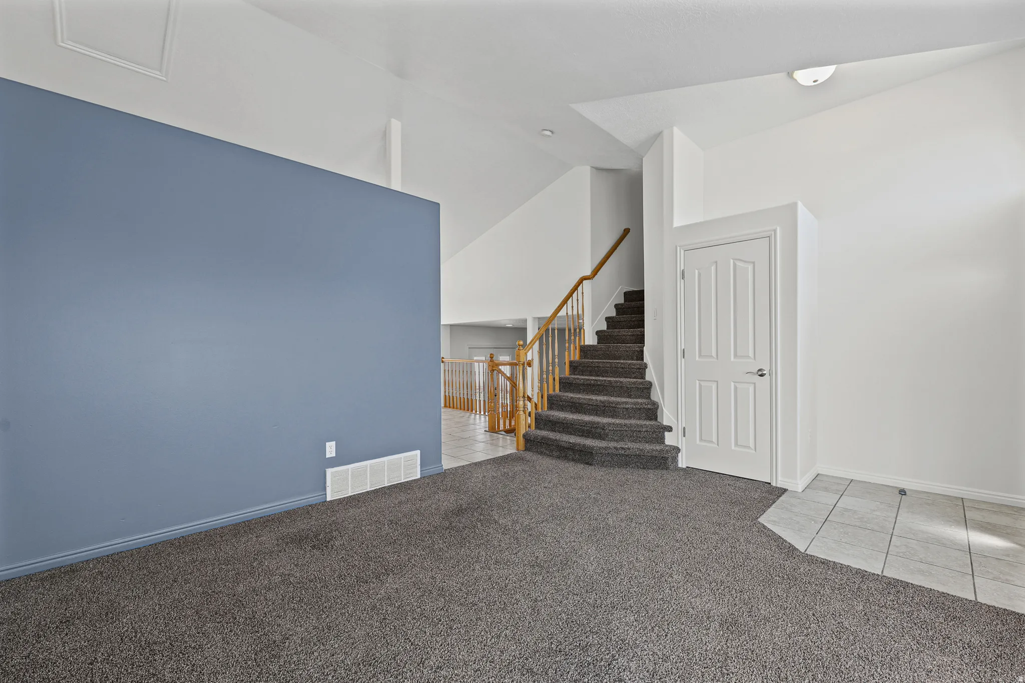 Vaulted ceilings above a carpeted room featuring a built-in staircase with wood railing, a solid core door, and tiled entry flooring