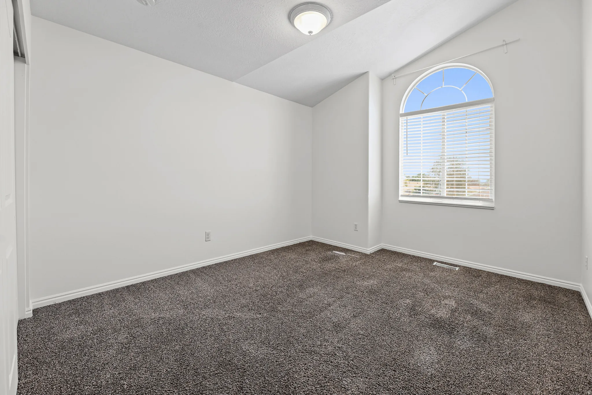 Room featuring a large arched window with blinds, vaulted ceiling, neutral wall paint, and dark gray carpet flooring