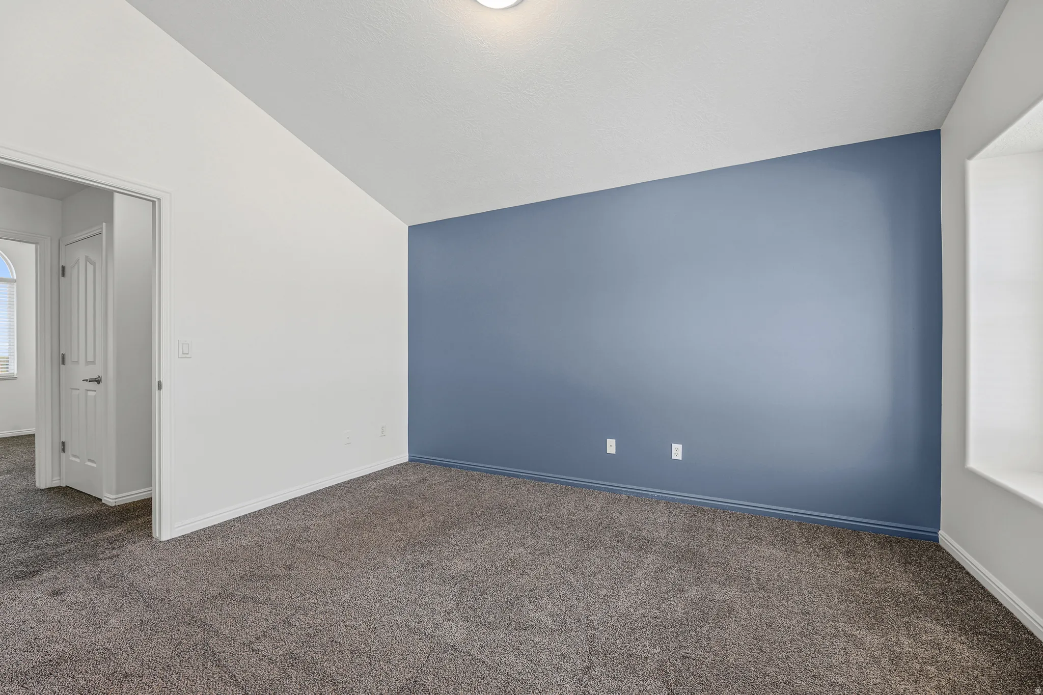 Spacious room featuring vaulted ceilings, plush carpet flooring, a distinctive accent wall, and a built-in window seat