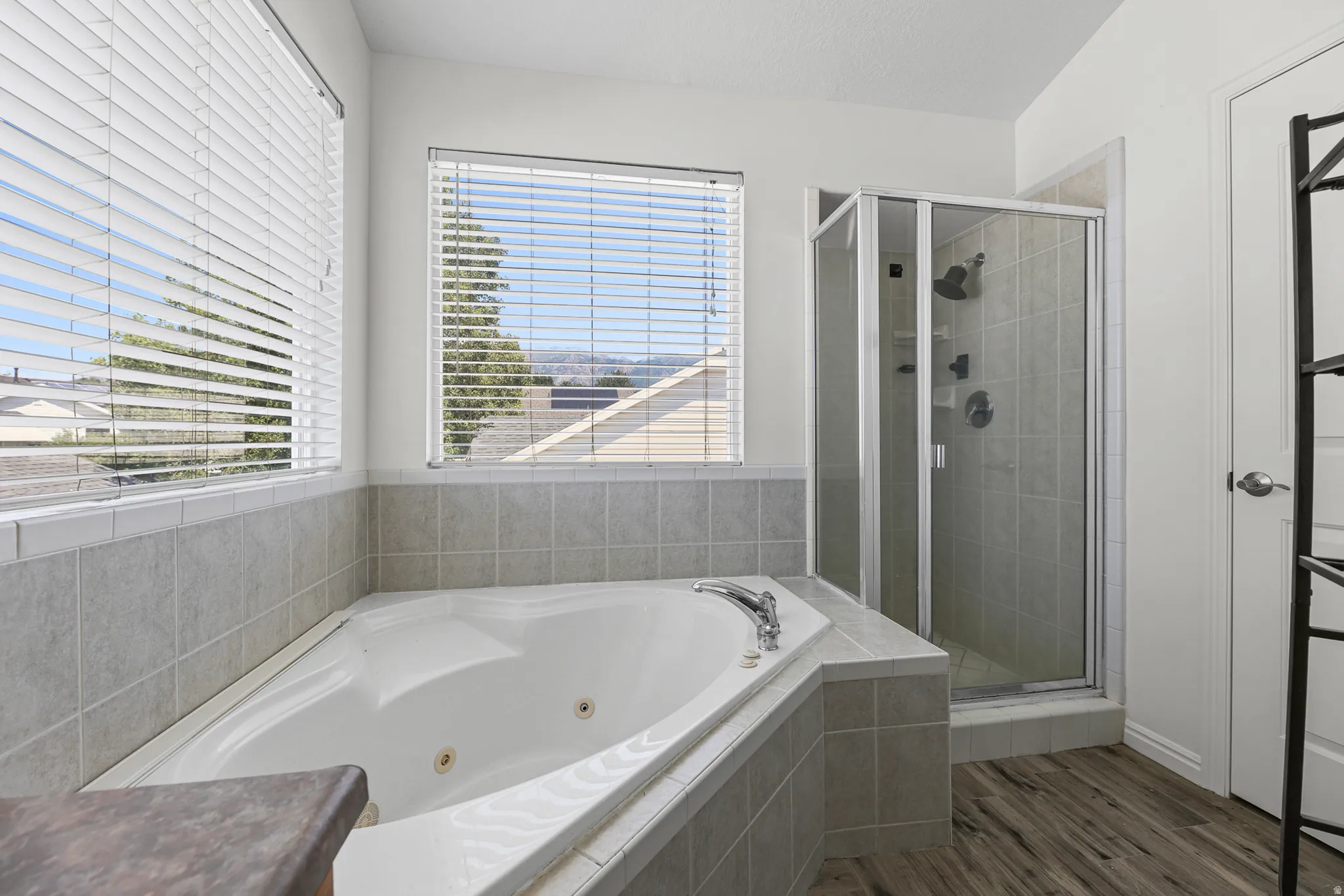 Soaking tub with jets, a tiled surround, two windows with blinds, an adjacent glass-enclosed shower, and wood-finish flooring