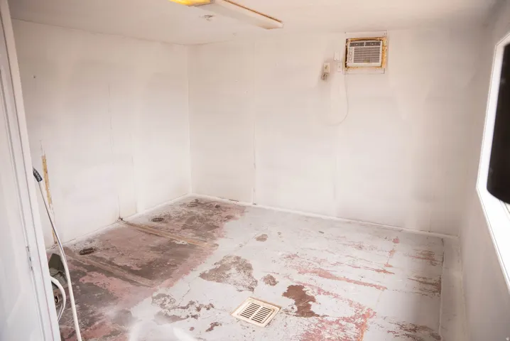 Unfinished interior space featuring a window-mounted air conditioning unit, a ceiling-mounted fluorescent light fixture, and a floor-level vent