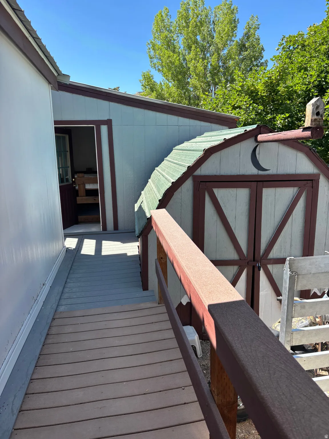 Exterior deck with wood-finish railing, leading to a light blue storage shed featuring a green gambrel roof and crescent moon detailing