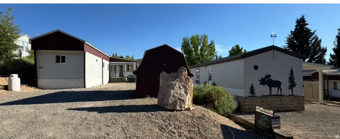 Gravel parking area fronting multiple structures, including a white-sided building with a dark roof and a separate white-sided building featuring a decorative moose and tree silhouette