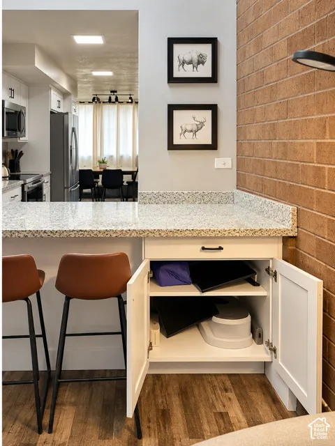 Kitchen peninsula featuring light-toned stone countertops, white shaker-style cabinetry with matte black hardware, and an exposed brick accent wall, finished with wood-finish flooring