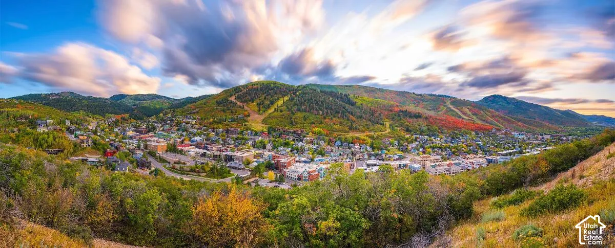Expansive mountain landscape featuring a valley town, surrounding hillsides with natural vegetation, and a wide sky with cloud formations
