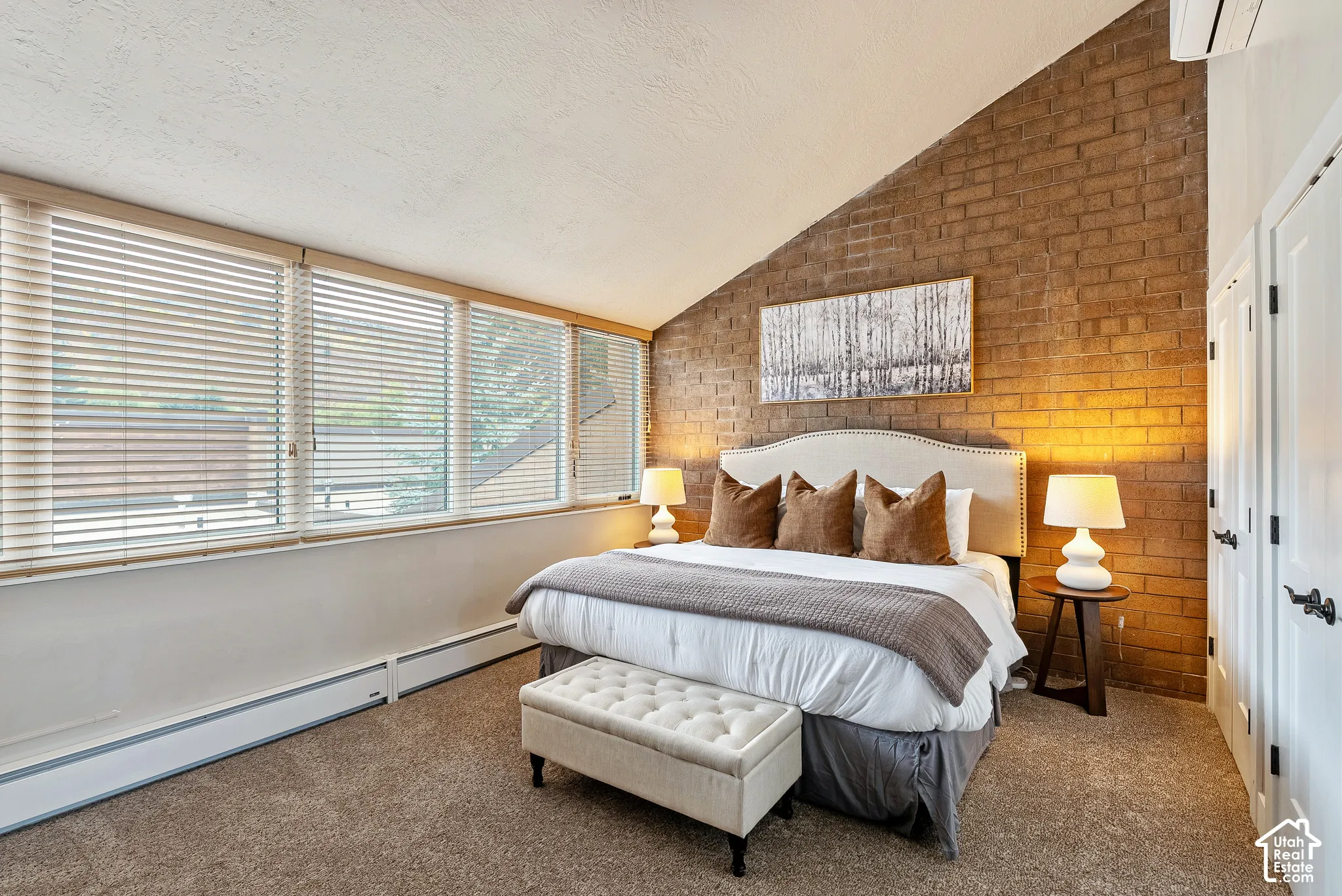 Vaulted ceiling bedroom featuring an exposed brick accent wall, a series of windows with horizontal blinds, built-in white cabinetry with dark hardware, and neutral-toned carpeting
