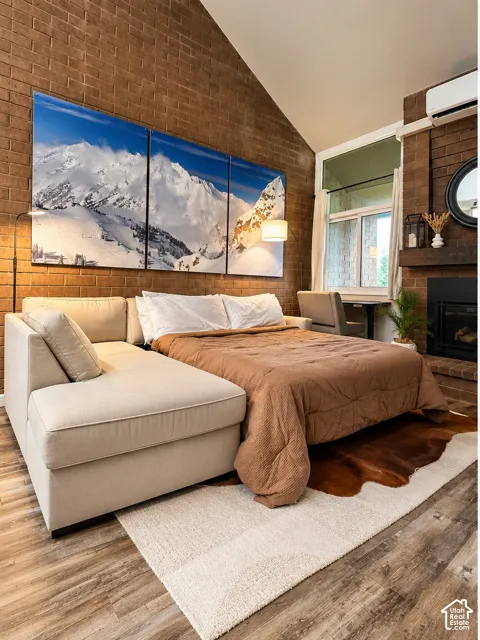 Vaulted ceiling featuring exposed brick walls, wood-finish flooring, a wall-mounted climate control unit, and a brick fireplace