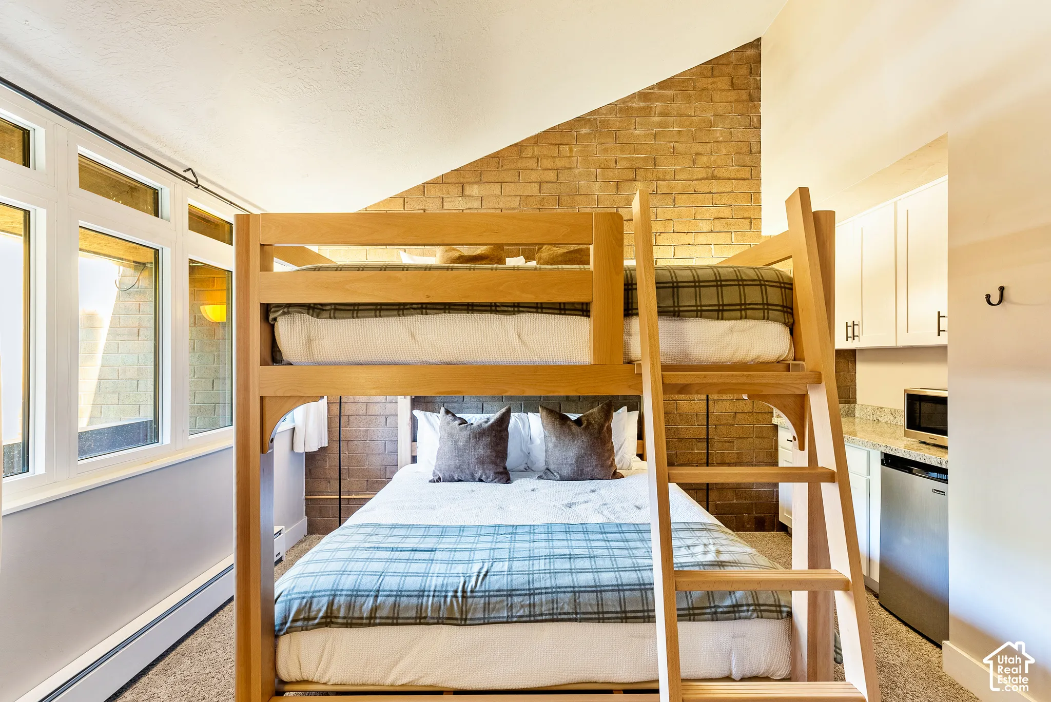 Room featuring a wood-finish bunk bed over a queen-size bed, brick accent wall, white-framed windows, and a built-in kitchenette with white cabinetry, a light-toned countertop, and a stainless steel mini-refrigerator