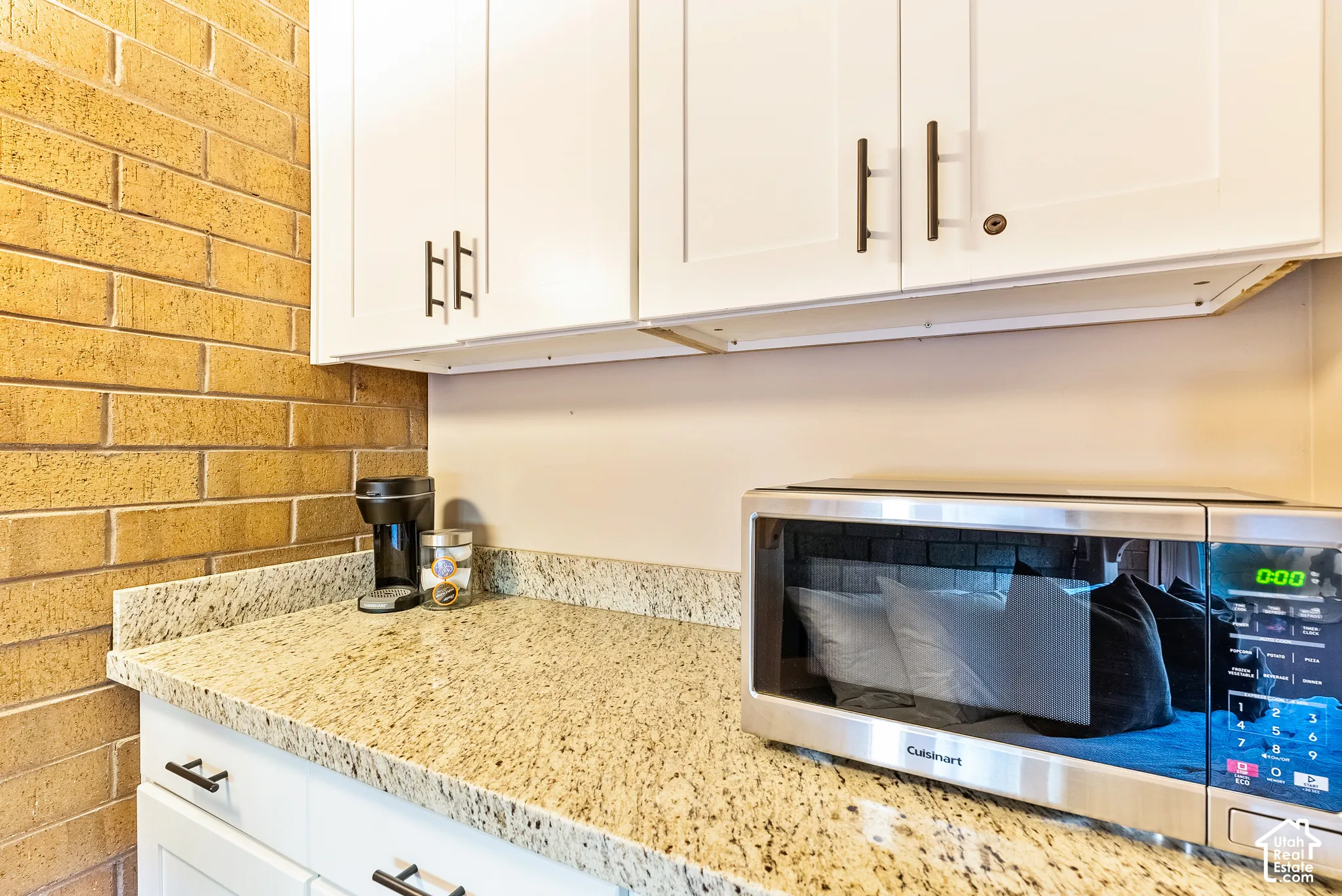 Kitchenette area featuring white shaker-style cabinetry with dark bronze hardware, a speckled granite countertop, and a brick accent wall