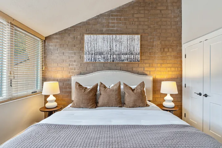 Bedroom featuring a striking exposed brick accent wall, white-paneled closet doors, and wood-finish window blinds