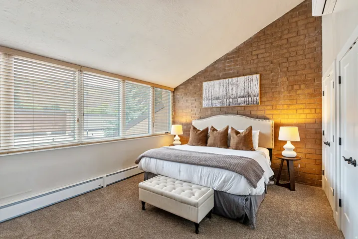 Vaulted ceiling bedroom featuring an exposed brick accent wall, a series of windows with horizontal blinds, built-in white cabinetry with dark hardware, and neutral-toned carpeting