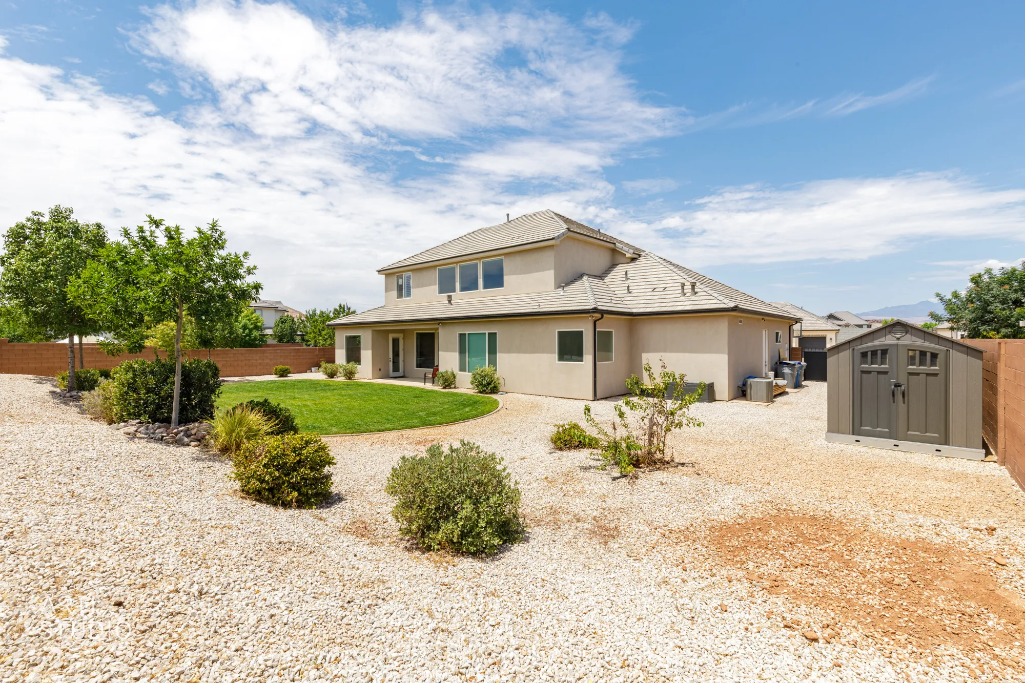 Rear exterior showcasing a neutral-toned stucco facade with multiple windows, a manicured lawn, gravel landscaping, and a freestanding storage shed
