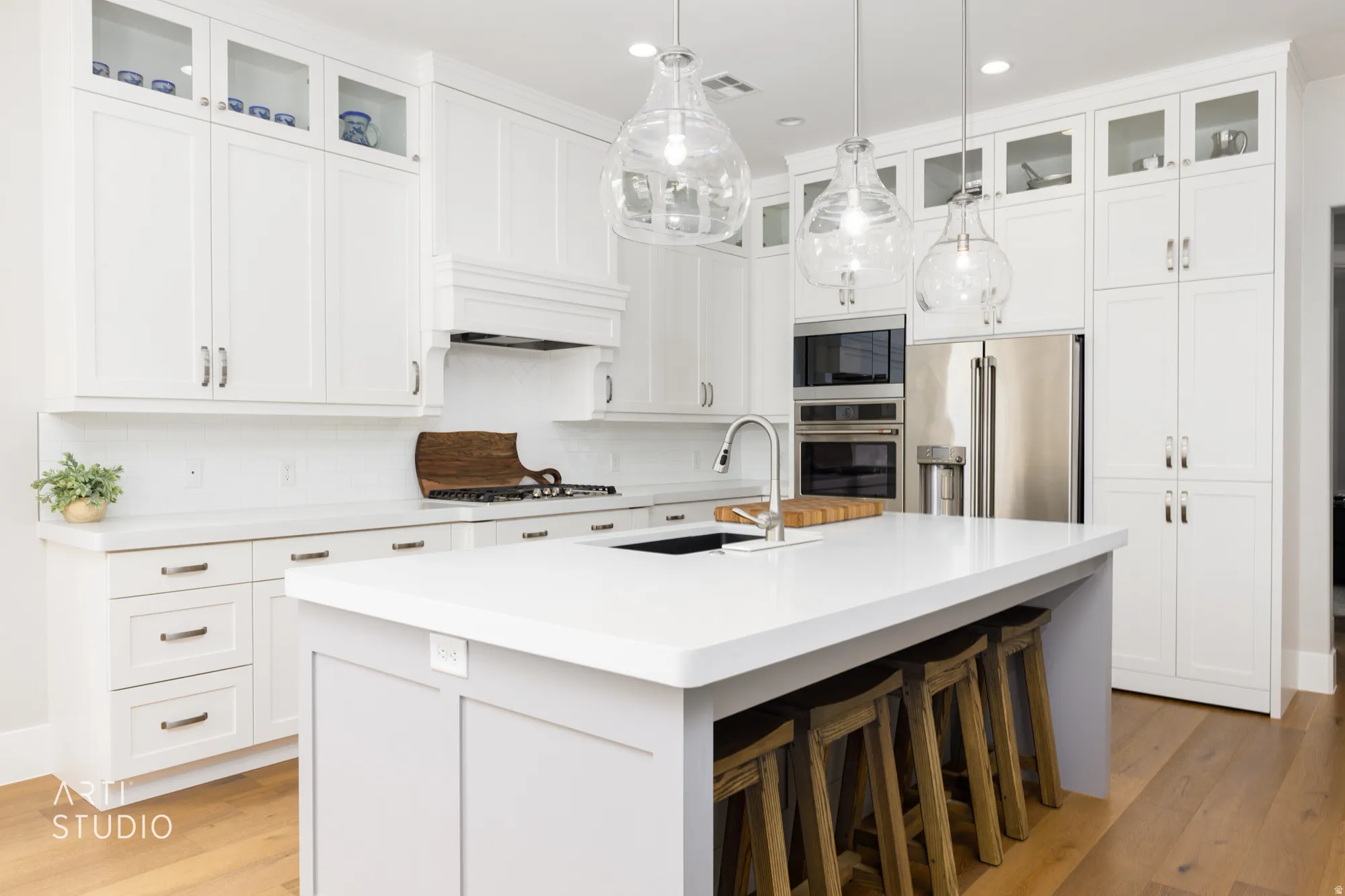 Bright white kitchen featuring extensive cabinetry with glass-front uppers, stainless steel appliances, a center island with countertop seating, and light wood-finish flooring