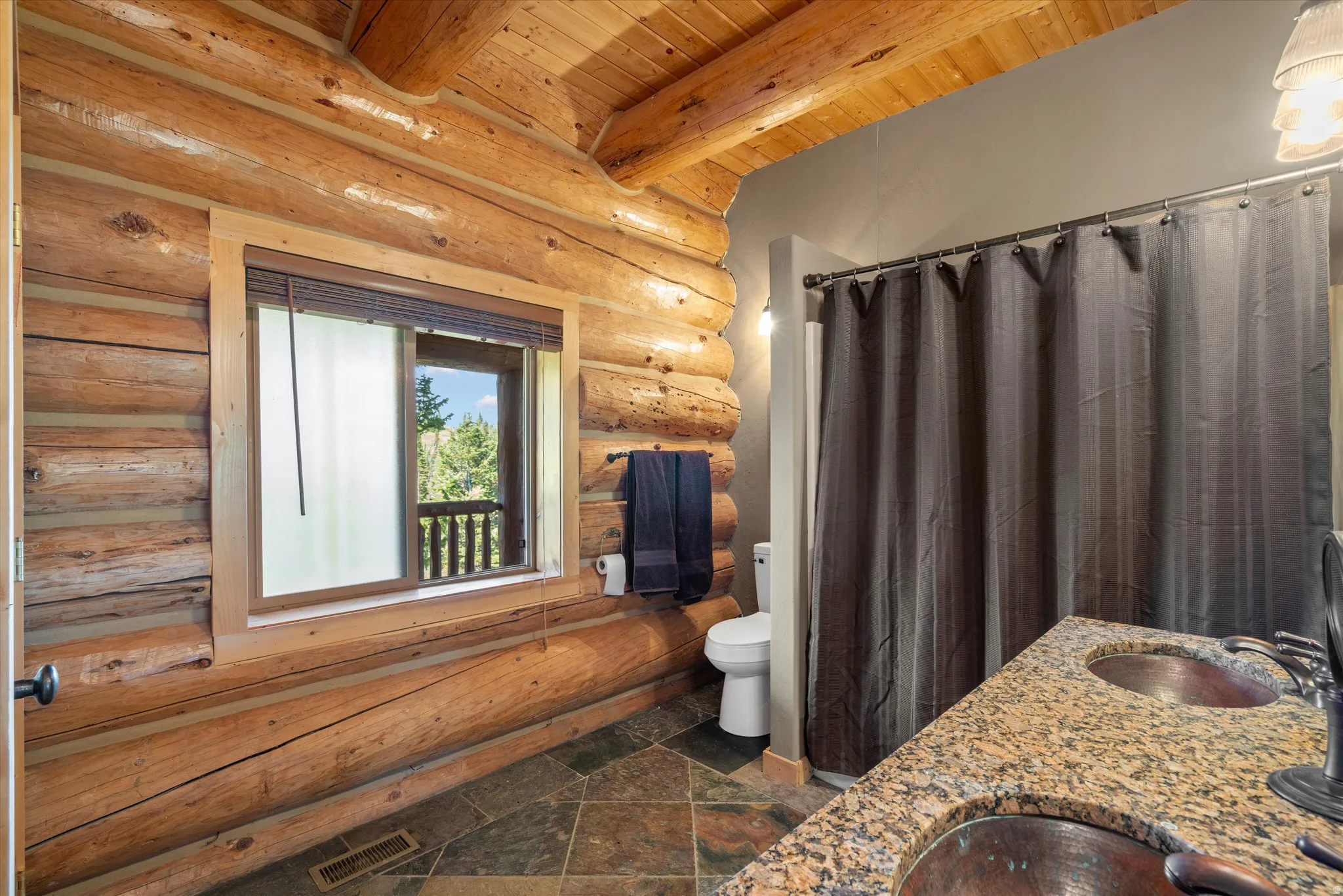 Bathroom featuring log wall construction, exposed wood beam ceiling, dual vessel sinks, stone countertop, and slate tile flooring