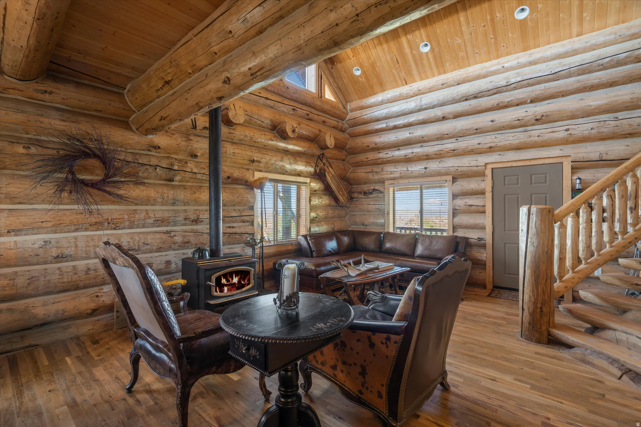 Spacious log cabin interior featuring a wood-burning stove with a visible flame, wood-finish flooring, exposed log walls, a vaulted wood-paneled ceiling with recessed lighting, and a handcrafted wooden staircase