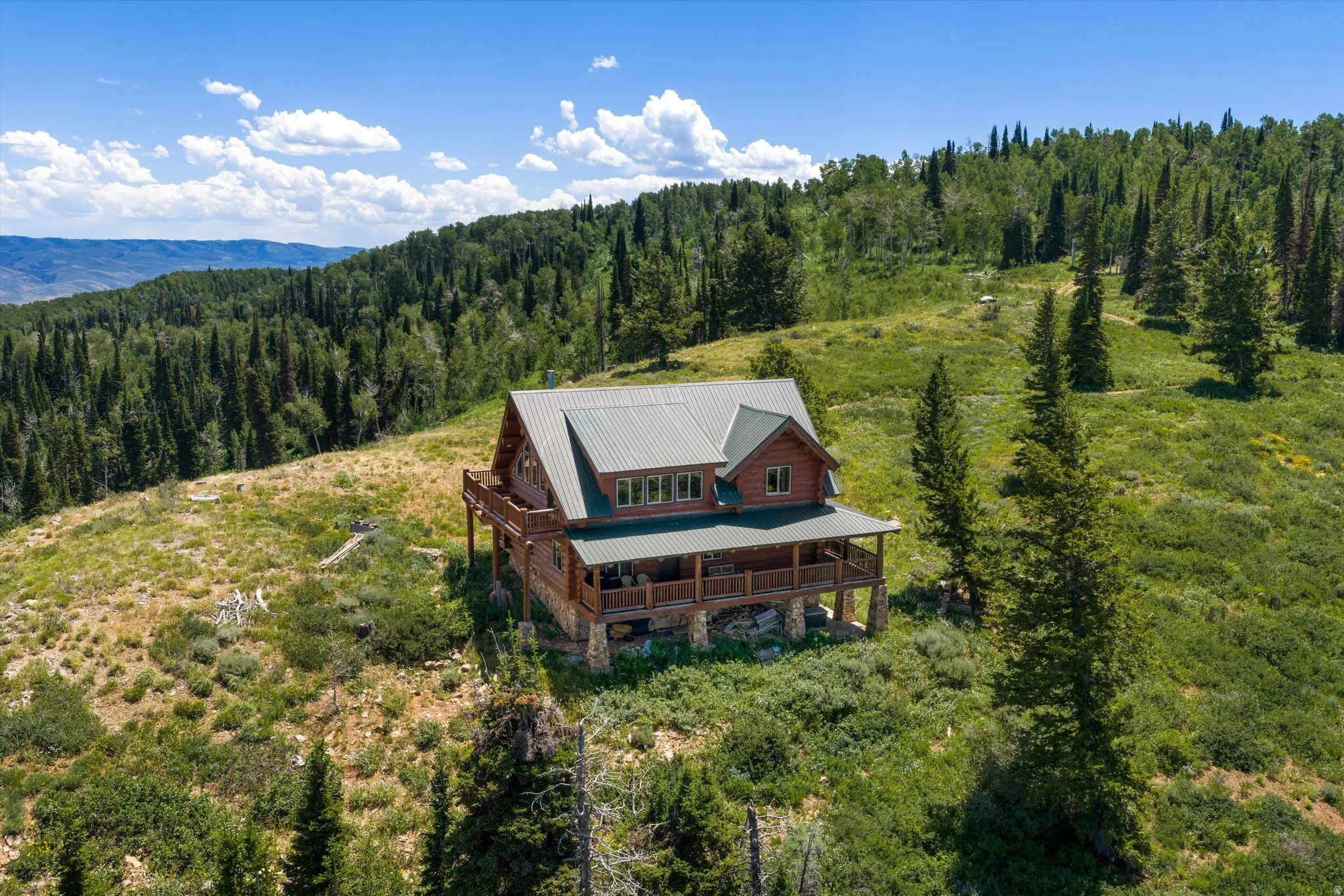 Rustic log cabin exterior with a green metal roof, featuring a full-length covered porch, an upper-level balcony, and a stone foundation