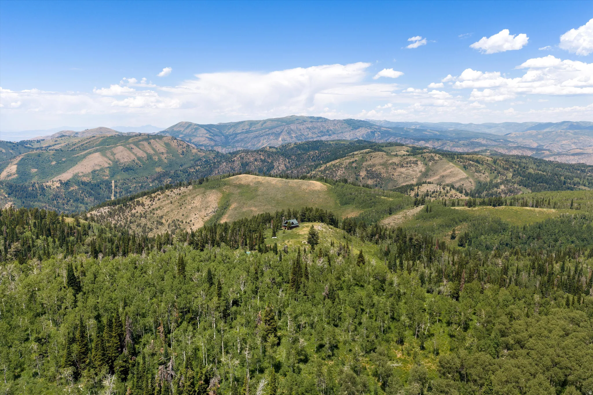 Expansive mountainous terrain featuring a secluded residence, dense evergreen forests, scattered deciduous trees, and undulating hillsides under a clear sky