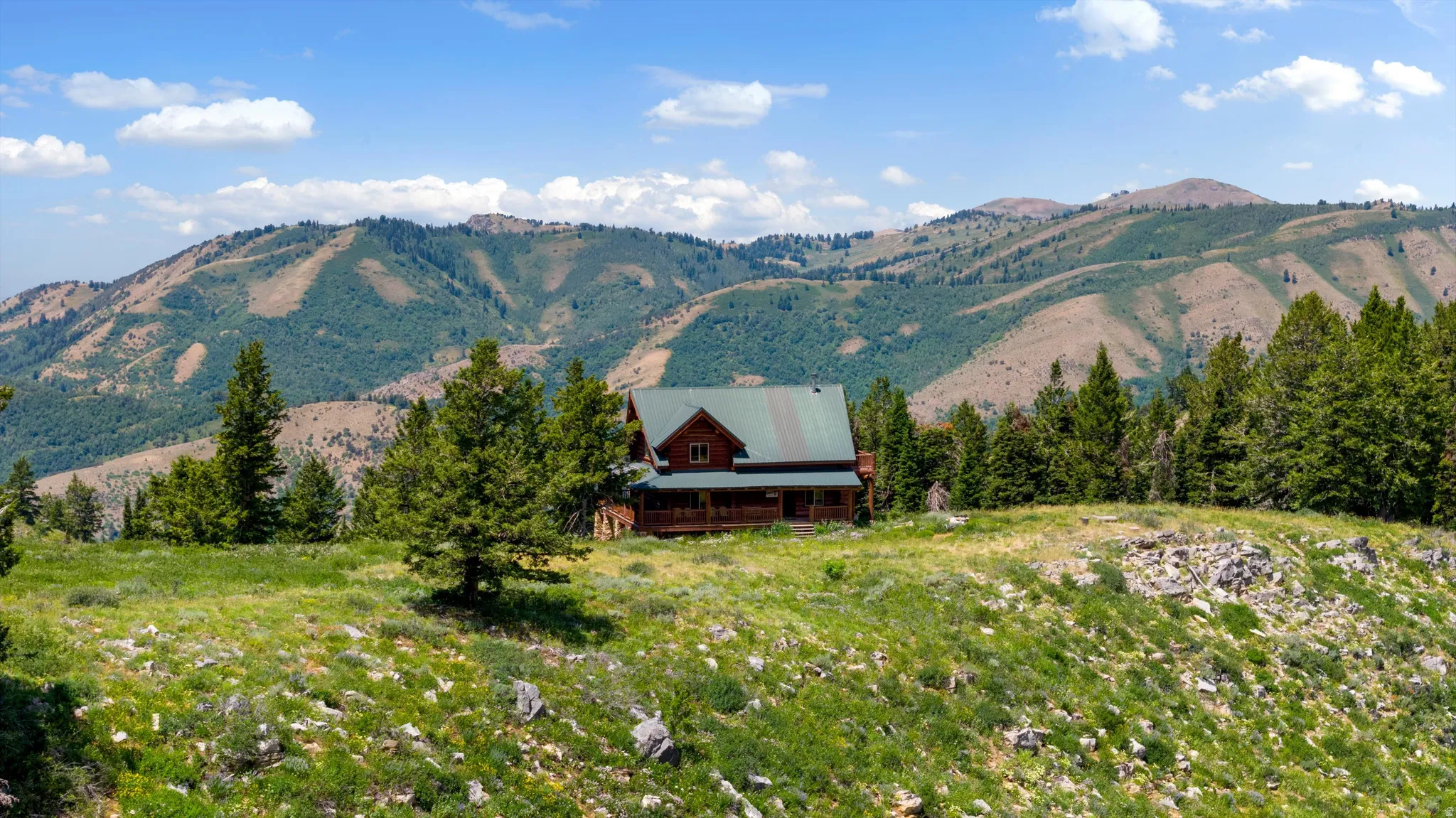 Elevated log home featuring a covered front porch and a green metal roof, set against a backdrop of mountainous terrain and mature evergreen trees