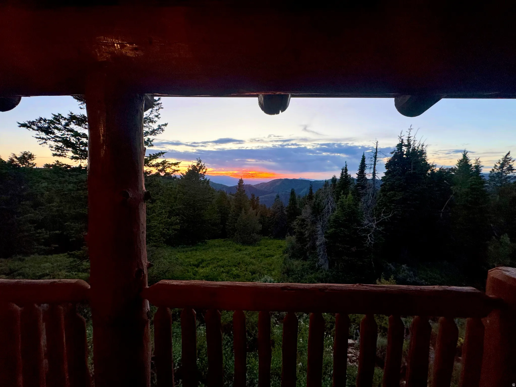 Rustic wood balcony featuring log railings and posts, framing a mountainous, tree-filled landscape