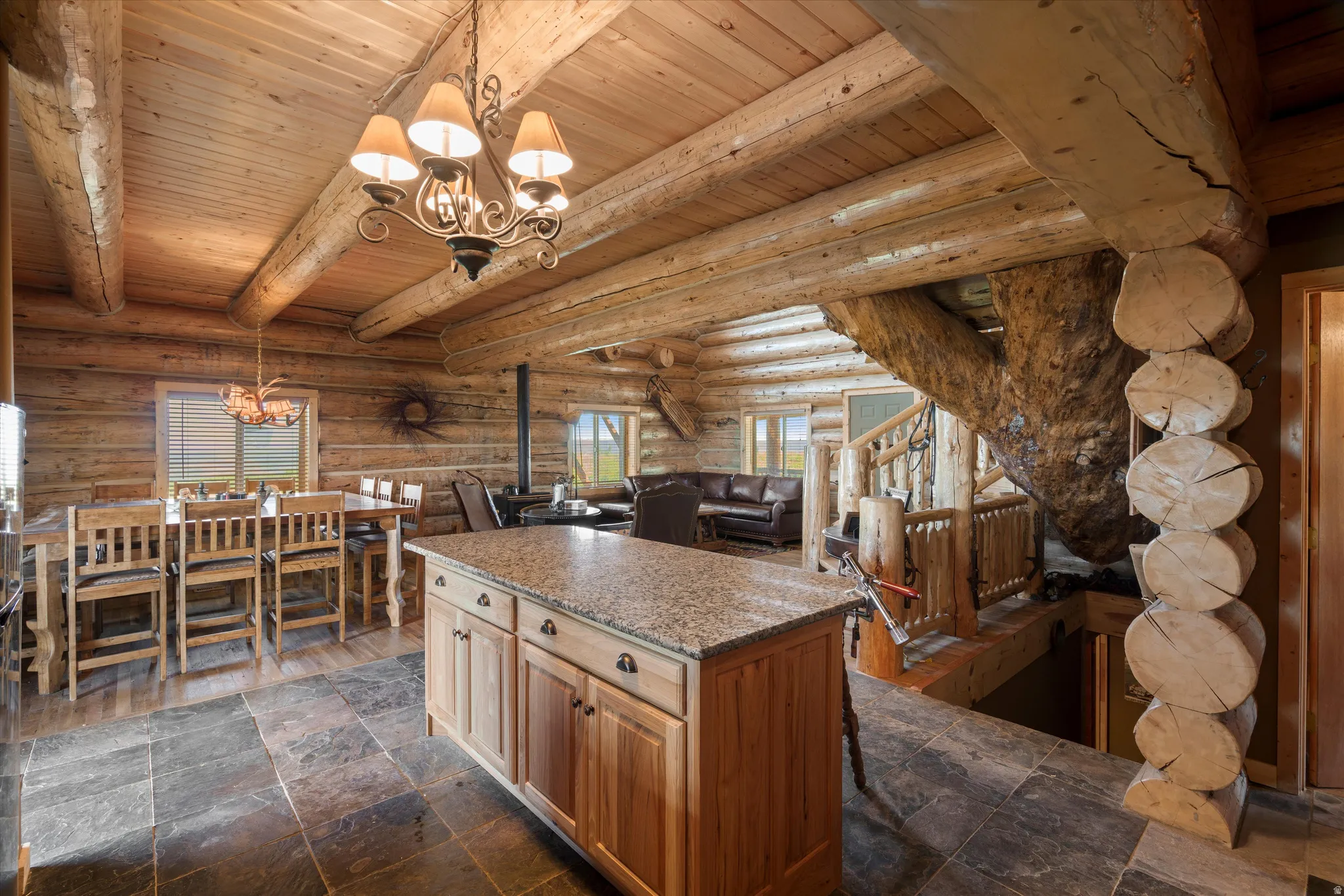 Open-concept interior featuring natural log walls and ceiling beams, a large wood island with a granite-style countertop, slate-style floor tiling, and a prominent log staircase