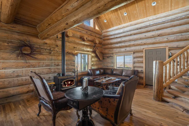 Spacious log cabin interior featuring a wood-burning stove with a visible flame, wood-finish flooring, exposed log walls, a vaulted wood-paneled ceiling with recessed lighting, and a handcrafted wooden staircase