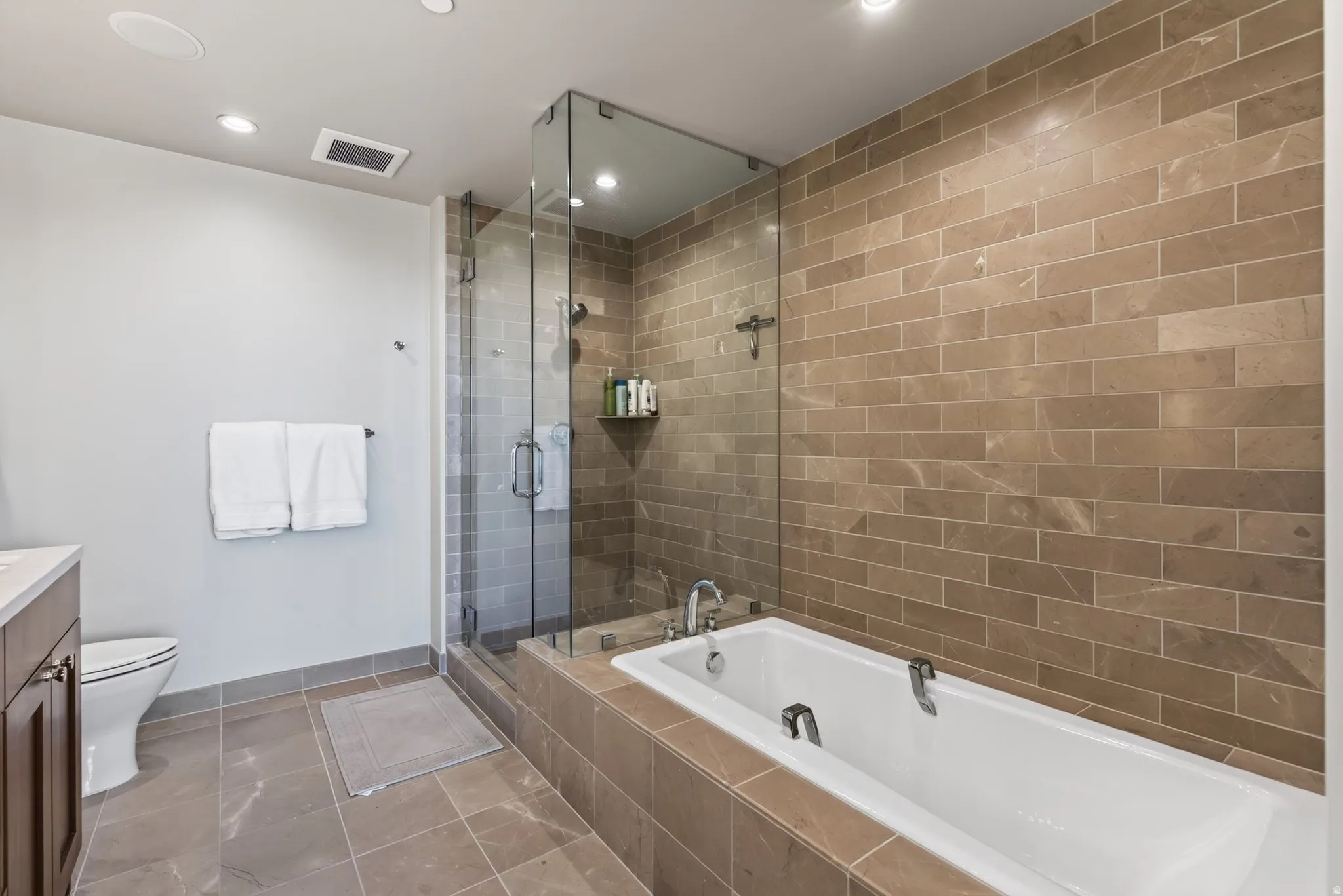 Spacious bathroom featuring a glass-enclosed shower, a built-in soaking tub, large format floor tiles, subway-style wall tiles, and recessed lighting