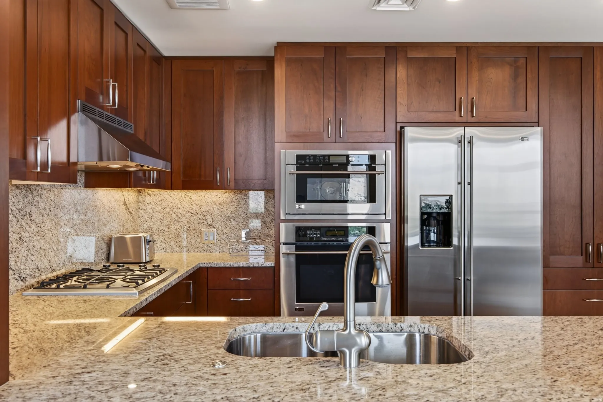Kitchen featuring rich wood cabinetry, stainless steel appliances including a double-door refrigerator and double wall ovens, a gas cooktop with a range hood, and speckled stone countertops with an undermount sink