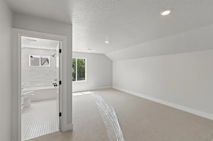 Recessed lighting, sloped ceiling, window with exterior greenery, neutral carpet, and a bathroom with a tiled tub surround and hexagonal floor tiles
