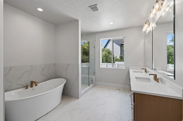 Freestanding soaking tub with gold-tone fixtures, walk-in shower with glass enclosure, dual vanity with undermount sinks, and large format marble-look tile flooring