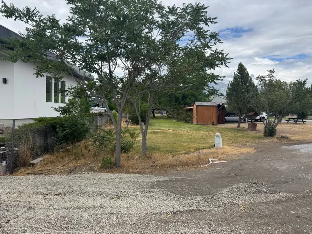 Expansive gravel and dirt lot featuring mature trees, a detached storage shed, a picnic table, and a built-in fire pit