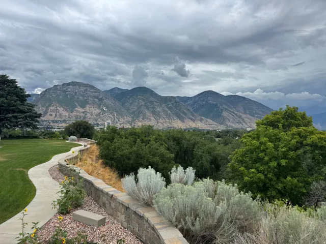 Expansive outdoor space featuring a winding concrete pathway, a stone retaining wall, mature landscaping, and distant mountain scenery