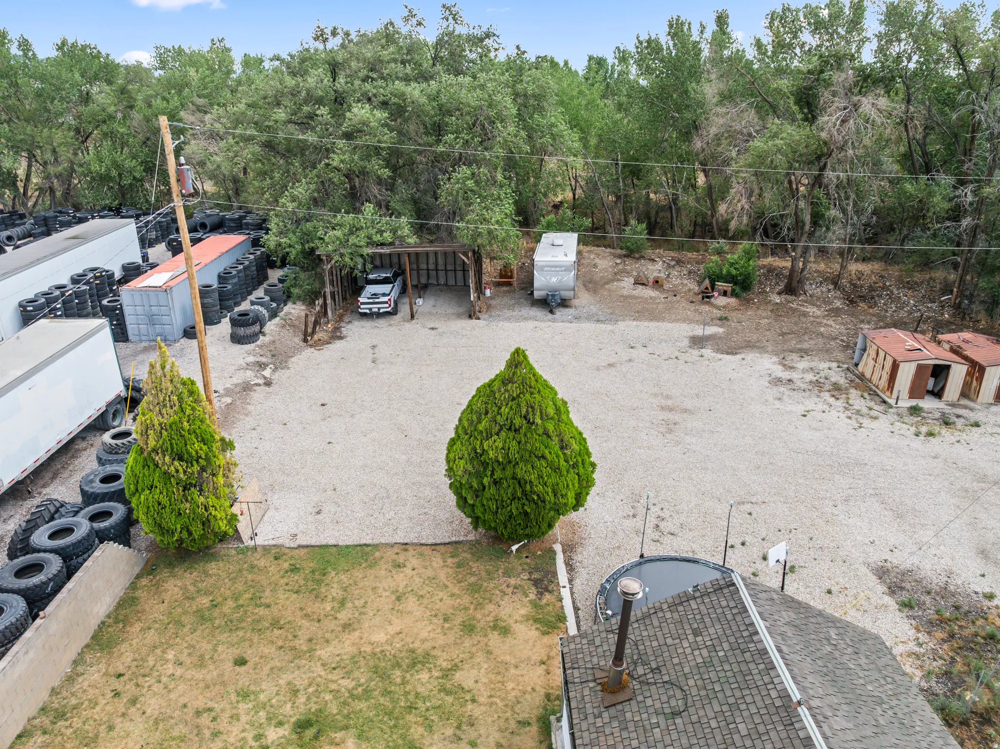 Expansive gravel lot featuring a covered parking structure, mature trees, and a partial view of a shingled roof