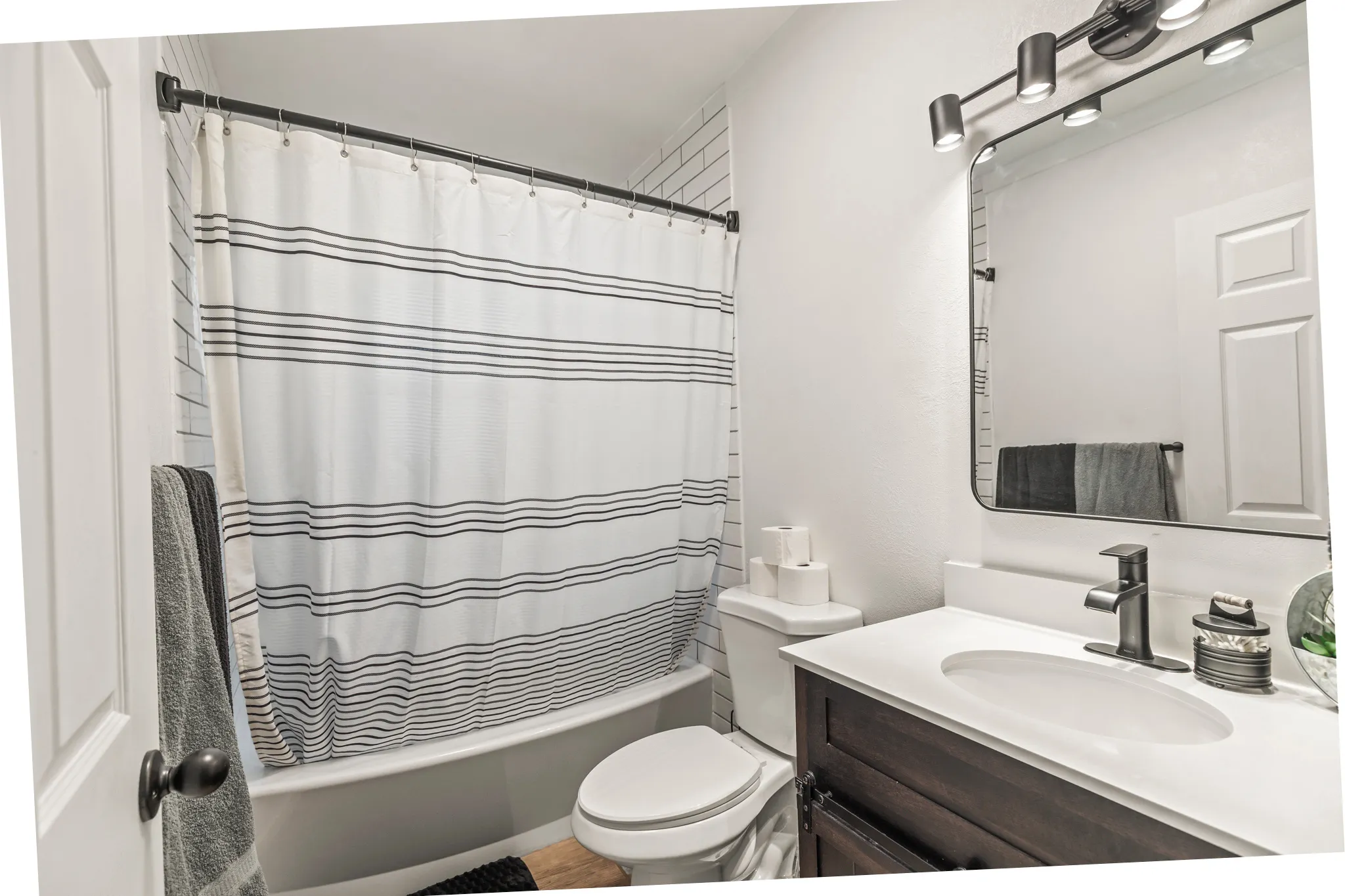 Bathroom featuring subway tile shower surround, a built-in tub, dark wood-finish vanity with a white countertop, and a contemporary black-framed mirror with integrated lighting