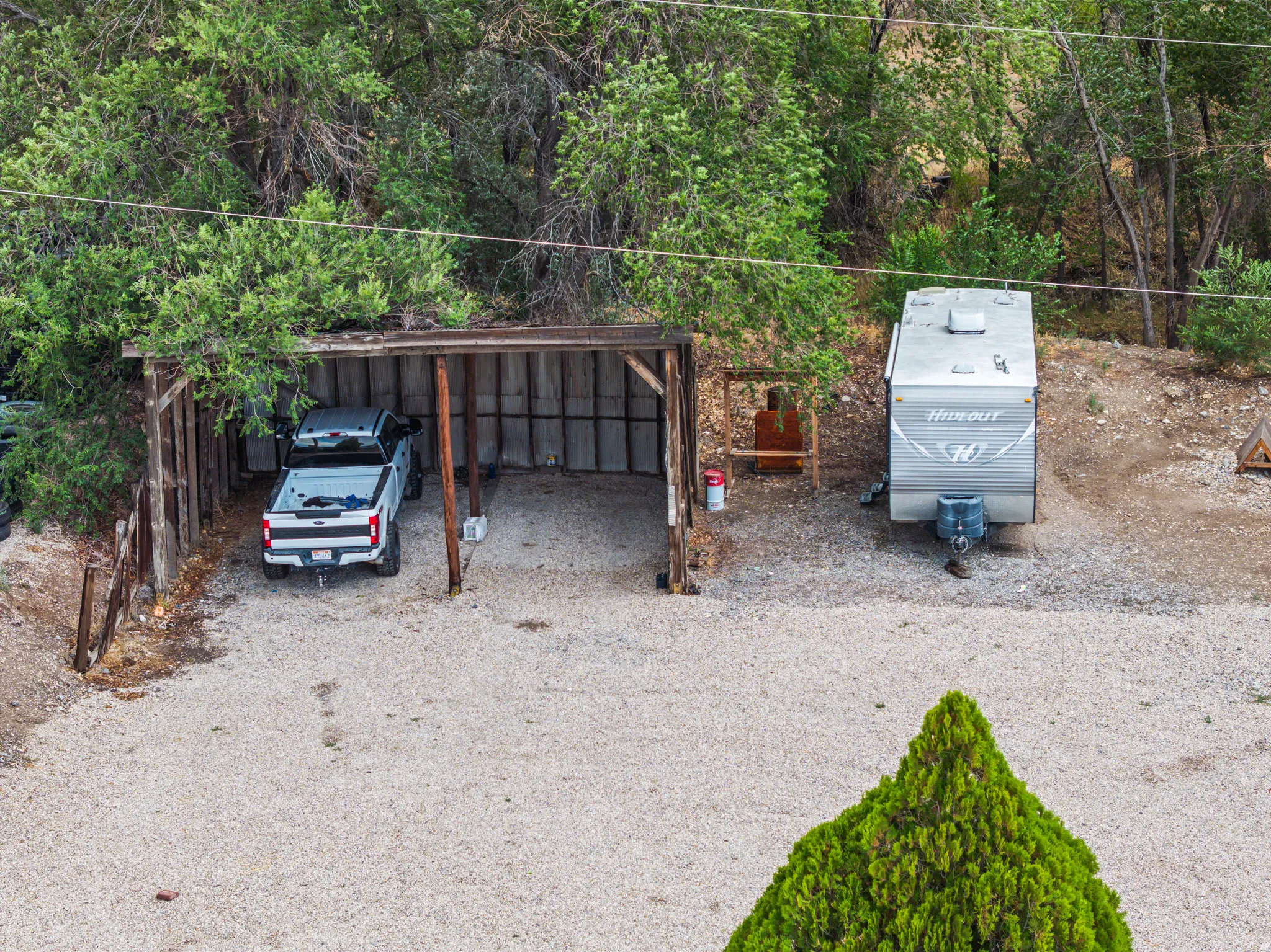 Gravel parking area featuring a wooden carport structure and surrounding mature trees
