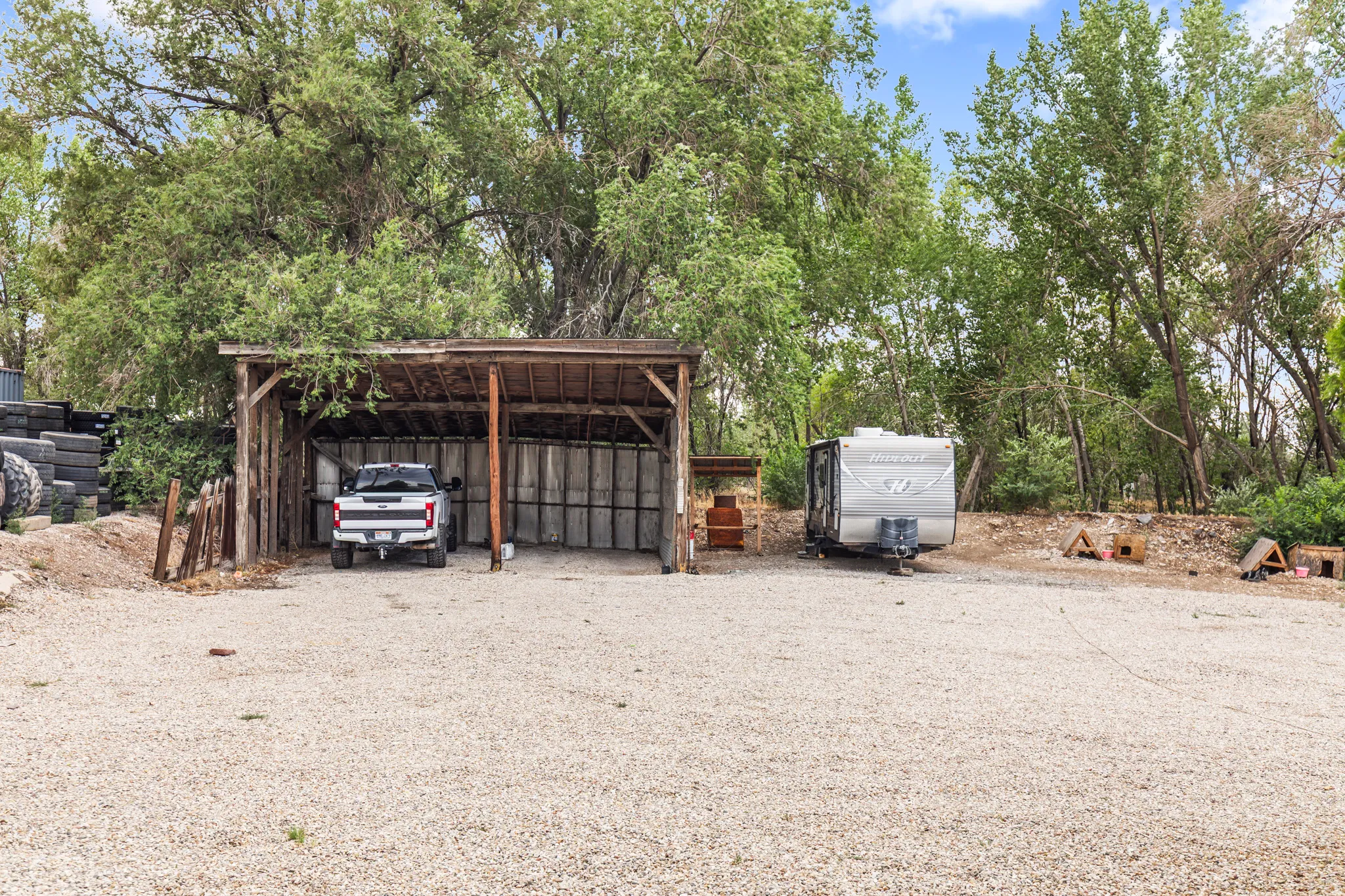 Gravel parking area featuring a rustic wood-framed carport, a covered storage structure, mature trees, and a partially wooded landscape