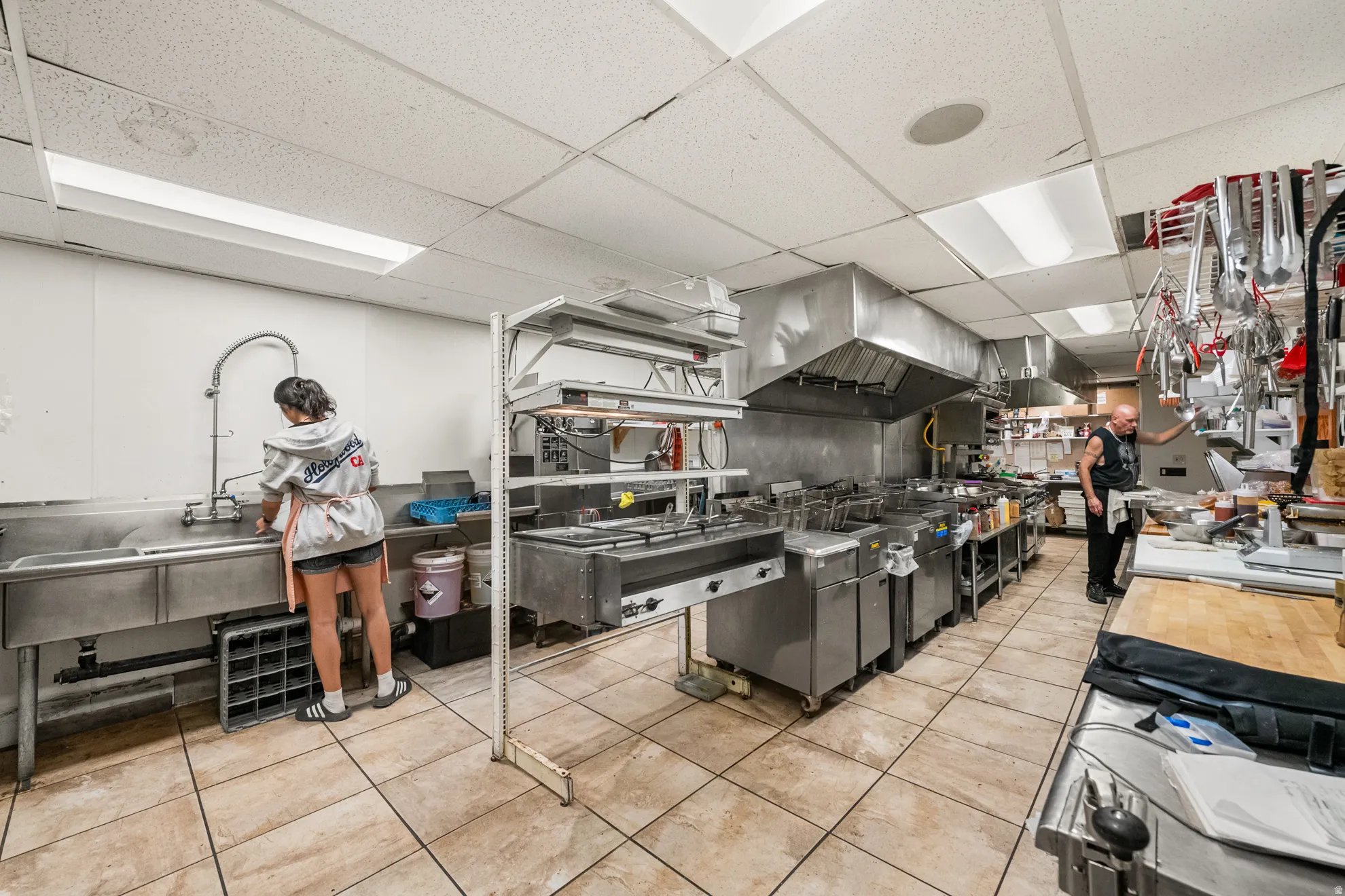 Commercial kitchen with multiple stainless steel workstations, a large multi-basin sink, extensive ventilation hoods, and tiled flooring