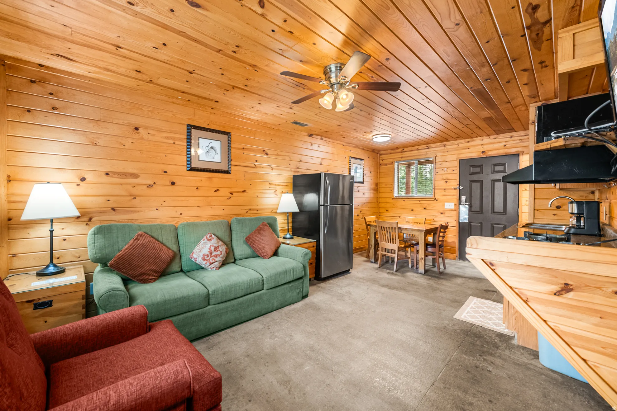 Open-concept living space featuring wood paneling on walls and ceiling, a ceiling fan, a full-size stainless steel refrigerator, and a compact kitchen counter with a built-in cooktop and range hood