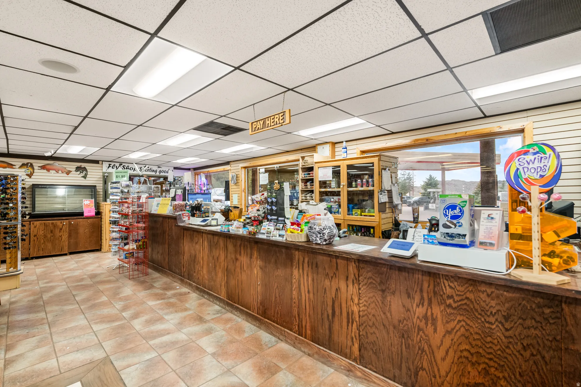 Retail space featuring a wood-finish sales counter, tiled flooring, drop ceiling with integrated lighting, and a large window