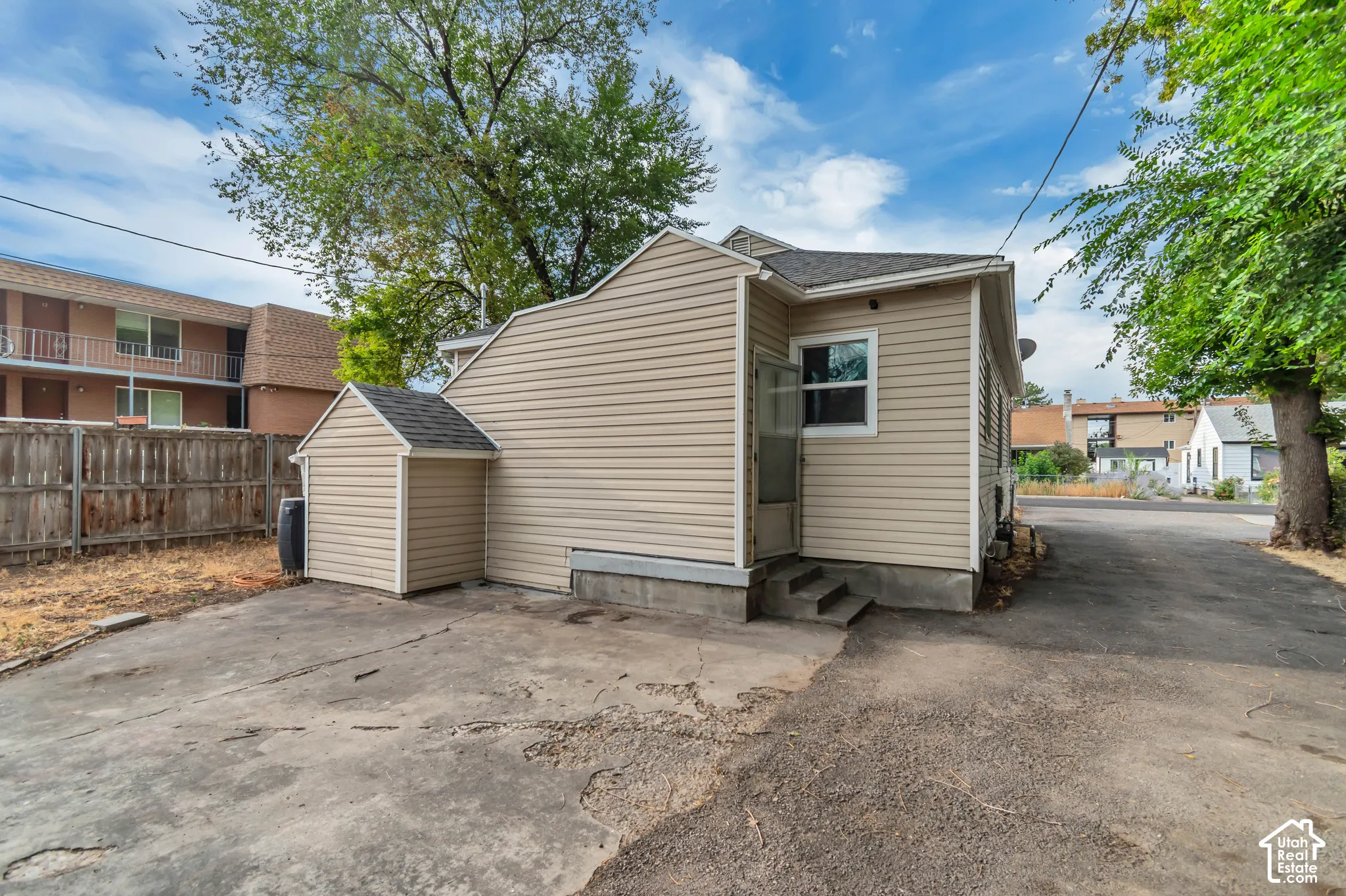 Exterior featuring beige horizontal siding, a detached storage shed with matching siding, concrete paving, and mature perimeter trees