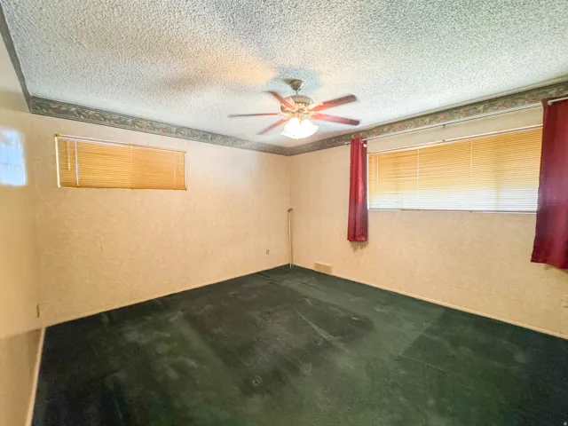 Room featuring textured wallpaper, green carpeting, a ceiling fan with integrated lighting, and two windows with horizontal blinds