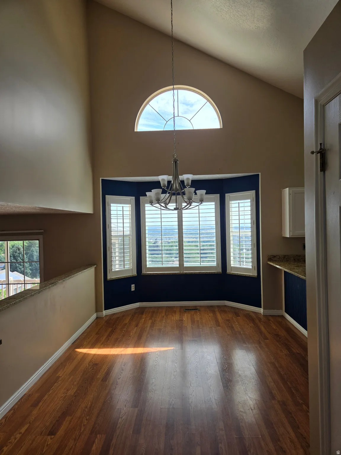 Vaulted ceiling with an arched transom window, wood-finish flooring, a bay window with plantation shutters, and a multi-arm chandelier