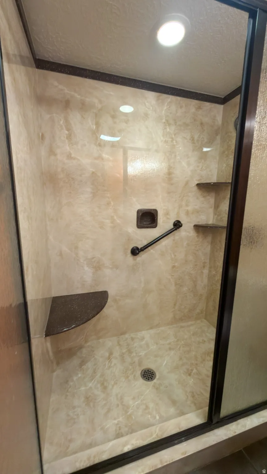Enclosed shower with marble-look wall and floor panels, featuring a built-in corner seat, integrated grab bar, recessed lighting, and a dark-framed glass door