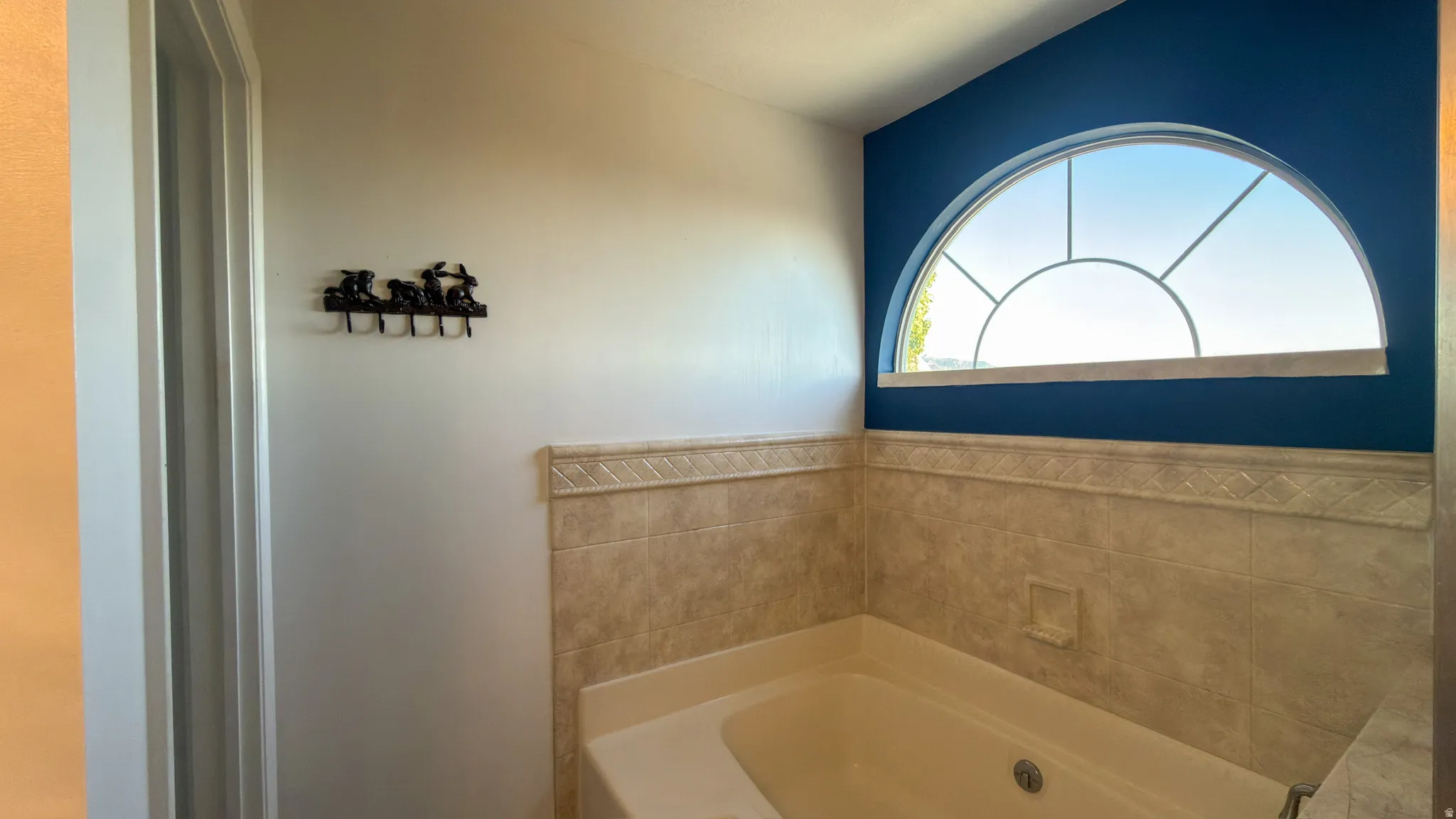 Soaking tub with tiled surround, contrasting accent wall, and an arched transom window