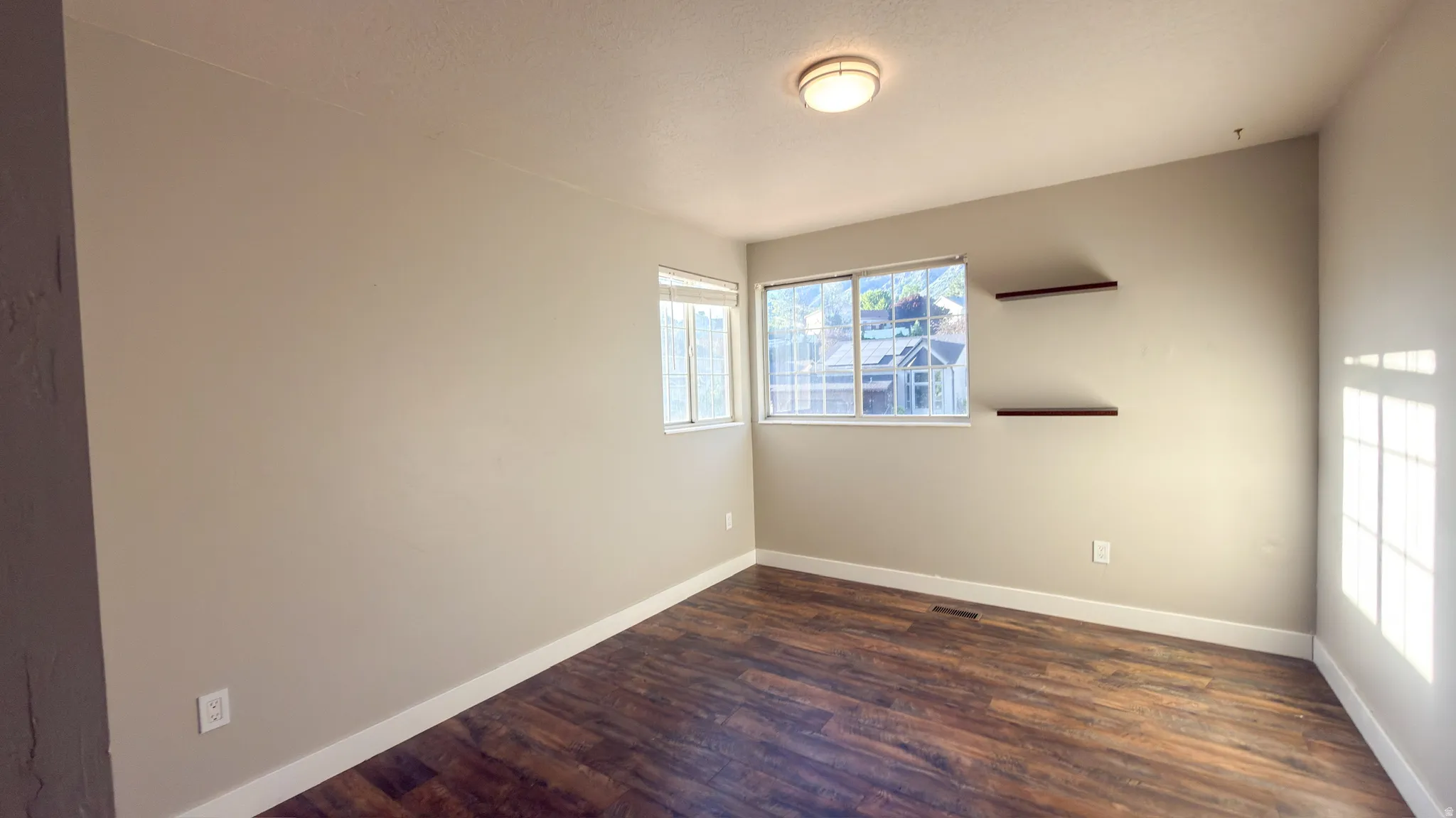Spacious room featuring wood-finish flooring, two windows, two built-in floating shelves, and a flush-mount ceiling light fixture