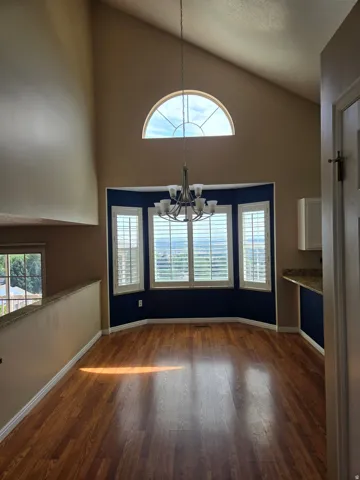 Vaulted ceiling with an arched transom window, wood-finish flooring, a bay window with plantation shutters, and a multi-arm chandelier