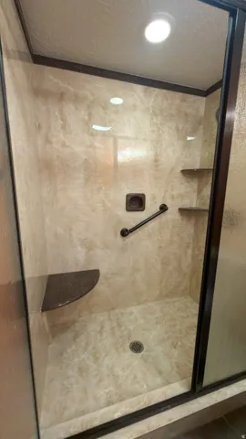 Enclosed shower with marble-look wall and floor panels, featuring a built-in corner seat, integrated grab bar, recessed lighting, and a dark-framed glass door
