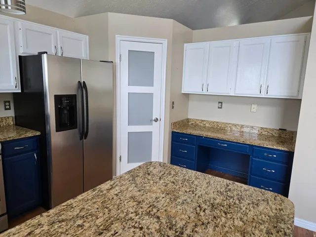 Kitchen featuring granite-style countertops, stainless steel side-by-side refrigerator, white upper cabinetry, blue lower cabinetry, and a pantry door with frosted glass panels