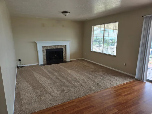 Spacious living area featuring a fireplace with stacked stone surround and white mantel, a large window with blinds, and a sliding glass door