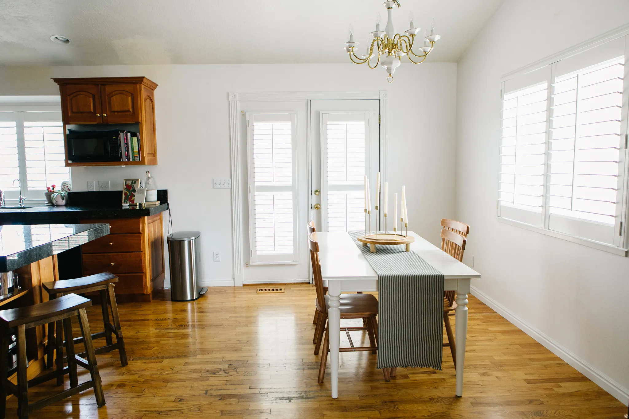 Kitchen and dining area featuring hardwood flooring, white plantation shutters, a white dining table with wood chairs, a kitchen island with dark countertops, and wood cabinetry