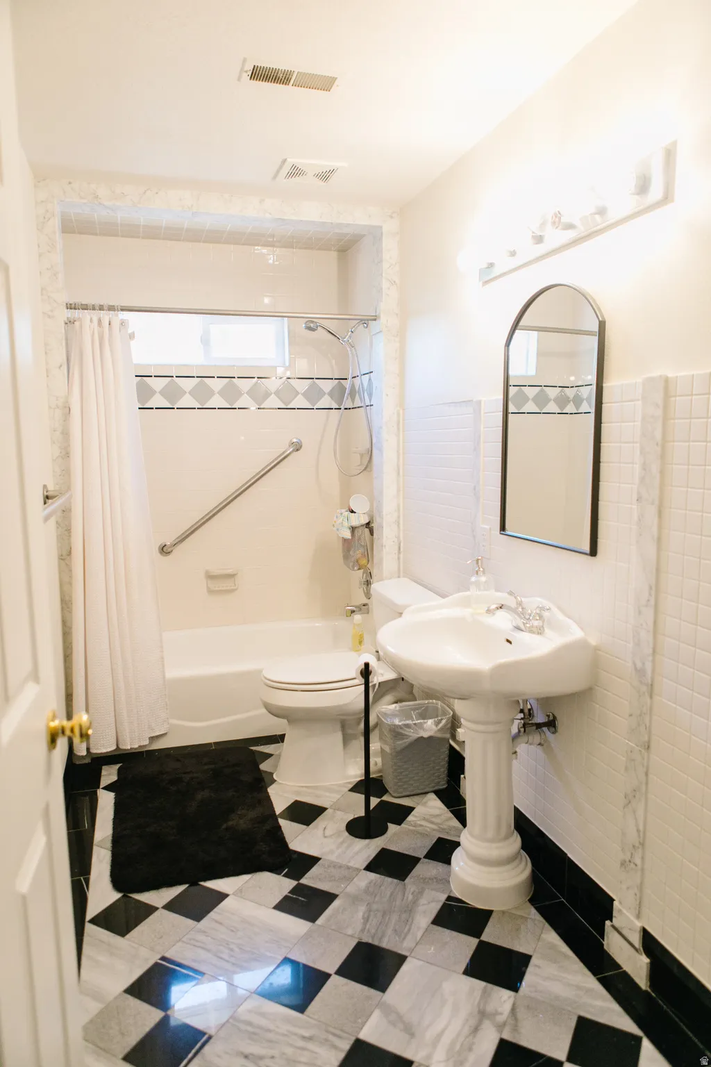 Bathroom featuring a black and white checkered marble floor, a pedestal sink, a bathtub with a tiled surround, a built-in grab bar, and a wall-mounted light fixture